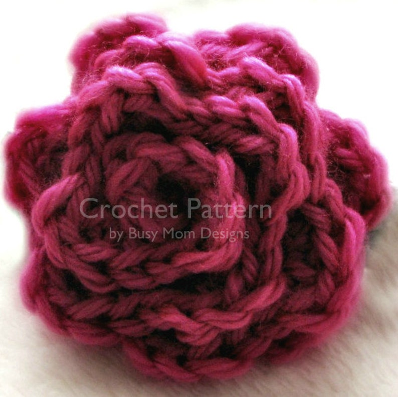 May include: A close-up of a crocheted rose made with a deep pink yarn. The rose is made with a simple stitch pattern and has a textured look.