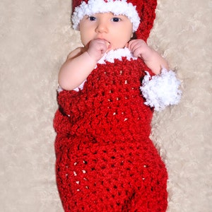 Puede incluir: Un bebé con un traje de Papá Noel de crochet rojo con ribete blanco. El bebé lleva un gorro de Papá Noel a juego con un pompón blanco.
