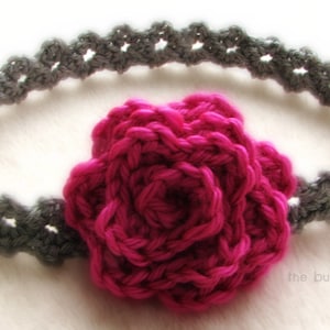 Op de afbeelding: Een grijs gehaakt hoofdband met een roze gehaakte bloem eraan bevestigd. Het hoofdband is gemaakt met een eenvoudige steek en de bloem is een ingewikkelder ontwerp.