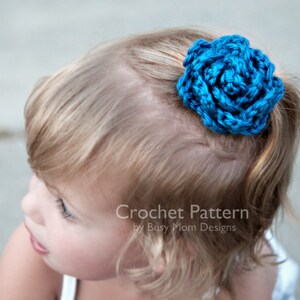 May include: A young girl with blonde hair wearing a blue crocheted flower hair clip. Crochet Pattern by Busy Mom Designs.