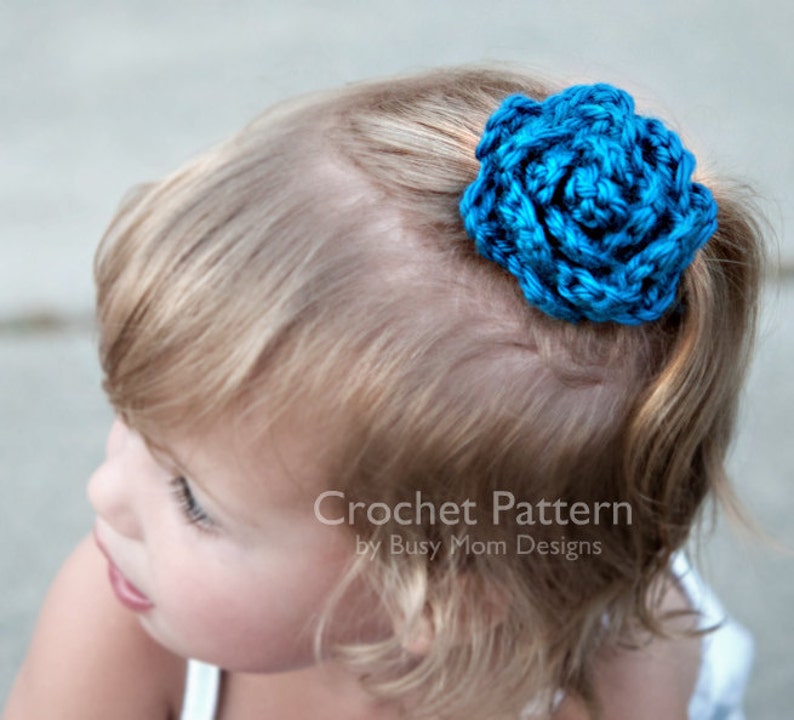 May include: A young girl with blonde hair wearing a blue crocheted flower hair clip. Crochet Pattern by Busy Mom Designs.