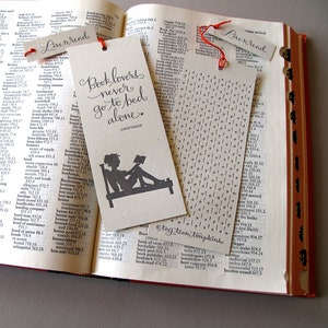 May include: A brown bookmark with the text "Book lovers never go to bed alone." in black script. The bookmark features a silhouette of a person reading a book in bed. The bookmark also includes the text "#tagteamtompkins" in black.