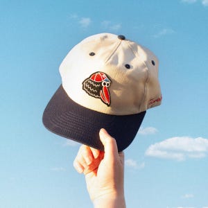 May include: A beige baseball cap with a navy blue brim. The cap features a patch with a cartoon bird design in red, black, and white. The word "Zurin" is written in red on the side of the cap.