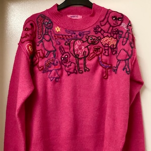 May include: A vibrant pink sweater with a crew neck and long sleeves. The sweater is adorned with embroidered cartoon characters in multiple colours across the chest and shoulders. The word "worn" is embroidered on the neckline.