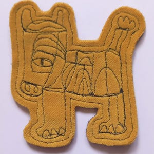 May include: A mustard yellow fabric patch shaped like a stylised four-legged animal. The design features black embroidered outlines that define the creature's head, body, legs, and tail, with simple geometric shapes forming the eyes and snout.