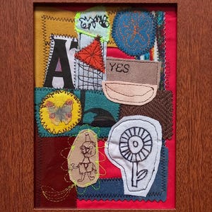 May include: A framed textile collage featuring a red background with various fabric patches including a black letter A, a yellow circle with a butterfly, a tan patch with the word YES, a white flower outline, and a hand-stitched figure in a brown wooden frame.