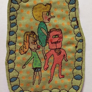May include: Embroidered patch with three cartoon figures on a light green background with orange polka dots. The patch has a blue and green border. The figures are in green, red, and orange. The patch is rectangular with rounded corners.