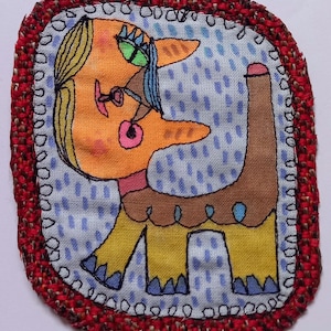 May include: A rectangular fabric patch featuring an embroidered illustration of a stylised orange cat with green eyes and yellow legs. The cat is set against a light blue background with dark blue rain-like dashes, all bordered by a thick red woven trim.