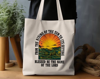 Può includere: Borsa tote in tela bianca con una grafica colorata di un tramonto e il testo "From the rising of the sun to its setting Blessed be the name of the Lord."