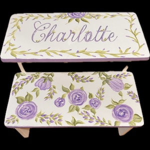 May include: A white wooden step stool with a purple and green floral design. The top step is painted with the name "Charlotte" in purple.