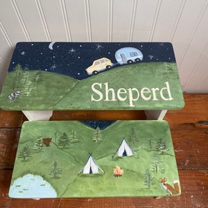 May include: A wooden children's step stool painted with a camping scene. The top of the stool features a night sky with stars, a yellow car pulling a blue camper, and the name "Shepherd". The bottom of the stool shows a green forest with two white tents, a campfire, and a fox.
