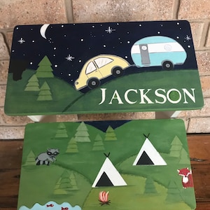 May include: A two-step wooden stool painted with a camping theme. The top step features a night sky with stars, a crescent moon, a yellow car pulling a blue camper, and the name "JACKSON". The bottom step shows a green forest with two white tents, a campfire, a raccoon, and a fox.