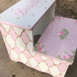 May include: A white and pink wooden step stool with a diamond pattern and floral design. The top step is painted pink with white polka dots and a floral design. The top step also has the name "Brielle" painted in pink cursive lettering.