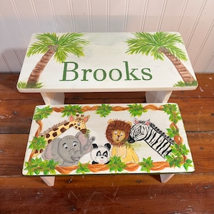 May include: White wooden step stool with a jungle theme. The top of the stool is painted with palm trees and the name "Brooks". The bottom of the stool is painted with a giraffe, elephant, panda, lion, and zebra.