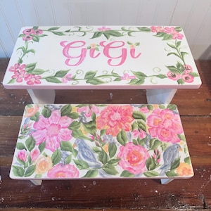 May include: A white wooden step stool with a floral design and the name "Gigi" painted on the top step. The bottom step features a floral pattern with pink and yellow flowers and blue birds.