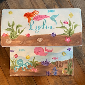 May include: Two rectangular, hand-painted wooden boxes with an underwater theme. The top box features a mermaid with red hair and the name "Lydia." The bottom box depicts a pink whale, octopus, and other sea creatures. The boxes are decorated with colorful fish, plants, and shells.