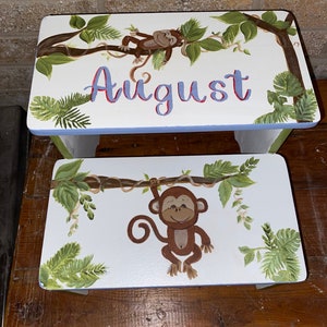 May include: Two white wooden step stools with a jungle theme. The top stool has the name "August" in blue and red lettering. Both stools feature a brown monkey, green leaves, and vines.