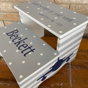 May include: A gray and white wooden step stool with a navy blue airplane silhouette on the side. The top of the stool is painted gray with white polka dots and the text "Guide my steps by your word Psalms 119:133". The bottom of the stool is painted with white and gray stripes. The name "Beckett" is painted in navy blue on the front of the stool.
