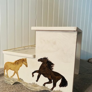 May include: A two-step wooden stool painted cream with horse illustrations. One horse is brown and rearing up, the other is tan and walking. The stool has a decorative edge and is suitable for children.