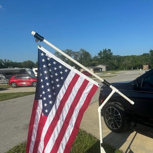 May include: American flag with red and white stripes and blue field of stars, mounted on a white pole. The flag is displayed outdoors, attached to a vehicle, with a clear blue sky in the background.