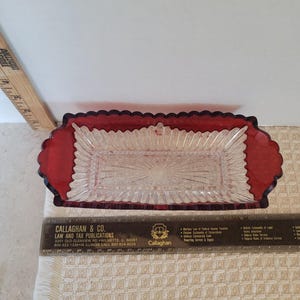 May include: A rectangular clear glass dish with a red border. The dish has a starburst pattern in the center and a scalloped edge. A ruler is placed near the dish.