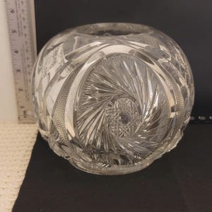 Puede incluir: Un cuenco de cristal transparente y redondo con intrincados patrones de vidrio tallado. El cuenco presenta un diseño en espiral en el centro y patrones florales en los bordes. El cuenco tiene unos 15 cm de diámetro.
