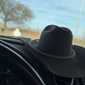 May include: A gray cowboy hat with a rope band rests on a car dashboard. The hat is the focal point, with a clear sky and a tree visible through the windshield. The steering wheel is in the foreground.