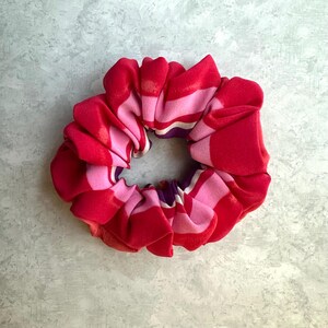 May include: A vibrant scrunchie in shades of red and pink, with a striped pattern. The fabric is gathered, creating a voluminous, circular shape. The scrunchie is a hair accessory.
