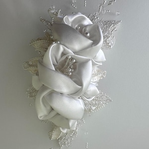 May include: A white satin flower arrangement with three large, rolled petals. The arrangement is accented with delicate lace, clear beads, and small pearl-like embellishments. The design is likely for a wedding or special occasion.
