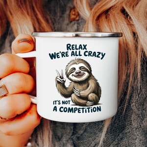 May include: White enamel mug with a silver rim featuring a cartoon sloth giving a peace sign. The mug has the text "RELAX WE'RE ALL CRAZY" and "IT'S NOT A COMPETITION" in dark blue.