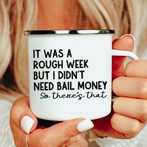 Op de afbeelding: Witte emaille mok met een zilveren rand en een wit handvat. De mok heeft de tekst "IT WAS A ROUGH WEEK BUT I DIDN'T NEED BAIL MONEY So there's that" in zwarte, handgeschreven letters.