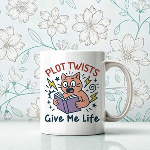 May include: White ceramic mug with the text "PLOT TWISTS GIVE ME LIFE" and a cartoon cat reading a book. The cat is orange with wide eyes and is surrounded by lightning bolts and stars. The mug has a curved handle.