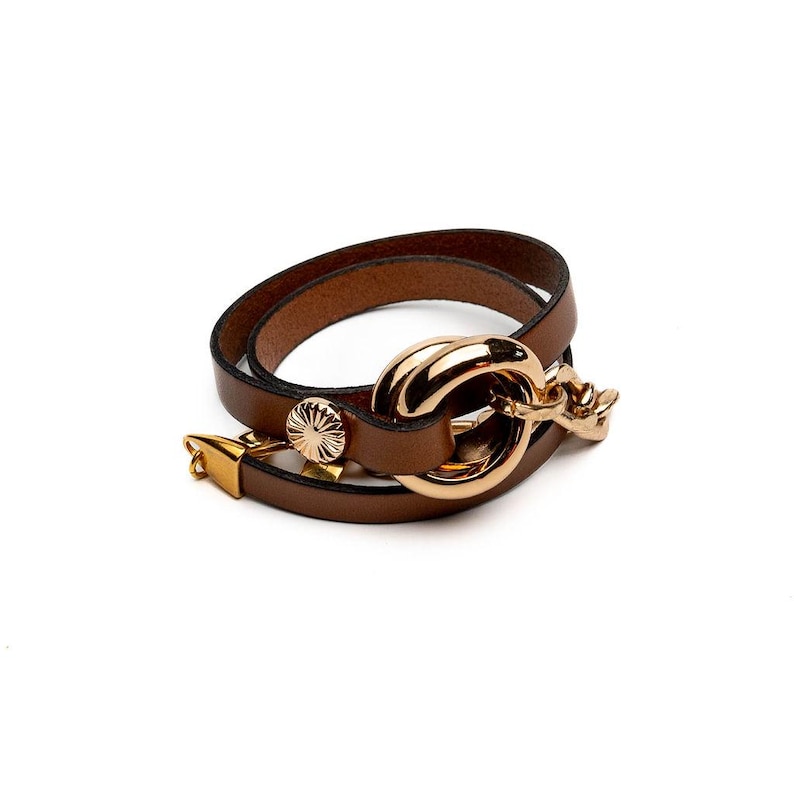 May include: Brown leather wrap bracelet with a gold-tone clasp and decorative accents. The bracelet features a double-wrap design with a large, circular gold-tone centerpiece and a smaller, decorative gold-tone clasp.