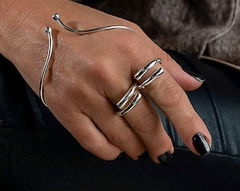May include: A silver cuff bracelet and a three-band ring. The cuff bracelet curves around the wrist with a small ball at each end. The ring has three parallel bands. The jewelry is displayed on a hand with black nail polish.