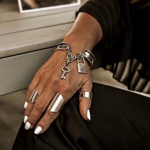 Può includere: Set di gioielli color argento con un braccialetto con un ciondolo a forma di lucchetto e chiave e diversi anelli. Gli anelli sono di vari design, tra cui una fascia larga e una fascia semplice. I gioielli sono indossati su una mano con smalto bianco.