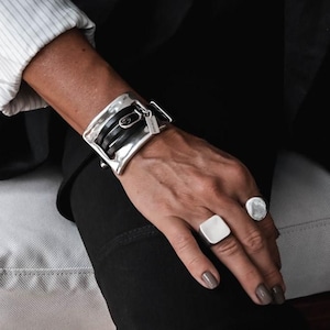 K&ouml;nnte beinhalten: Ein silberfarbenes Armband mit Schnallendesign, kombiniert mit einem quadratischen und einem runden Silberring. Das Armband hat schwarze Lederriemen. Der Schmuck wird an einer Hand getragen, mit einem schwarzen Outfit im Hintergrund.