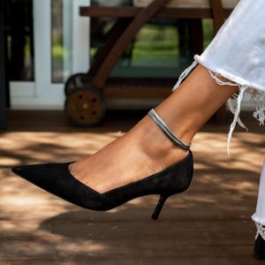 Puede incluir: Zapatos de tacón negros de ante con punta y tacón pequeño. Se lleva una pulsera de tobillo plateada y gris en el tobillo. El fondo incluye una terraza de madera y un entorno exterior borroso.