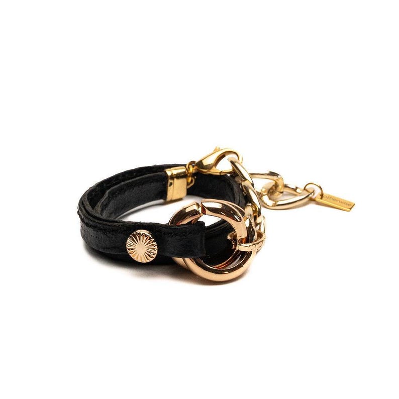 Puede incluir: Una pulsera de cuero negro con detalles met&aacute;licos dorados. La pulsera presenta un cierre dorado, un bot&oacute;n decorativo dorado y una cadena con una peque&ntilde;a etiqueta. La pulsera est&aacute; sobre un fondo blanco.