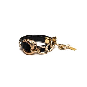 Puede incluir: Una pulsera de cuero negro con eslabones y herrajes dorados. La pulsera presenta un cierre dorado y una peque&ntilde;a etiqueta dorada. La pulsera est&aacute; sobre un fondo blanco.