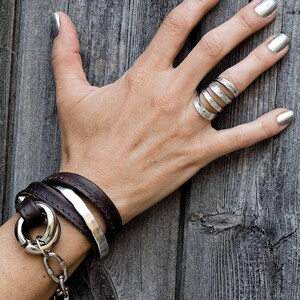 Puede incluir: Una mano con esmalte de u&ntilde;as plateado muestra un anillo de varias bandas y una pulsera envolvente. La pulsera presenta una correa de cuero marr&oacute;n oscuro, una banda de plata martillada y un gran anillo plateado con una cadena y un dije de cuerno.