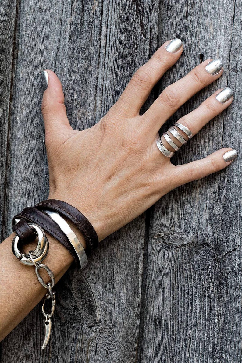 Puede incluir: Una mano con esmalte de u&ntilde;as plateado muestra un anillo de varias bandas y una pulsera envolvente. La pulsera presenta una correa de cuero marr&oacute;n oscuro, una banda de plata martillada y un gran anillo plateado con una cadena y un dije de cuerno.