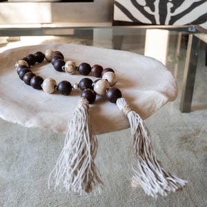 May include: A decorative beaded garland with dark brown, cream, and silver beads, and two cream-colored tassels. The garland is displayed on a white, organic-shaped dish, suggesting a home decor item.