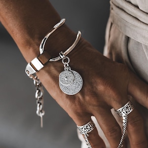 Könnte beinhalten: Ein silberfarbenes Armband mit einer glatten, geschwungenen Stange, einem braunen Lederakzent und einem hängenden Anhänger mit kreisförmigen Gravuren. Zwei passende silberfarbene Ringe mit filigranen Ausschnitten sind durch eine Kette verbunden.