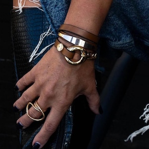May include: A collection of gold and brown bracelets and a gold ring. The bracelets include a gold cuff, a brown leather wrap, and a gold chain bracelet. The ring is a unique open design. The jewelry is worn on a wrist.