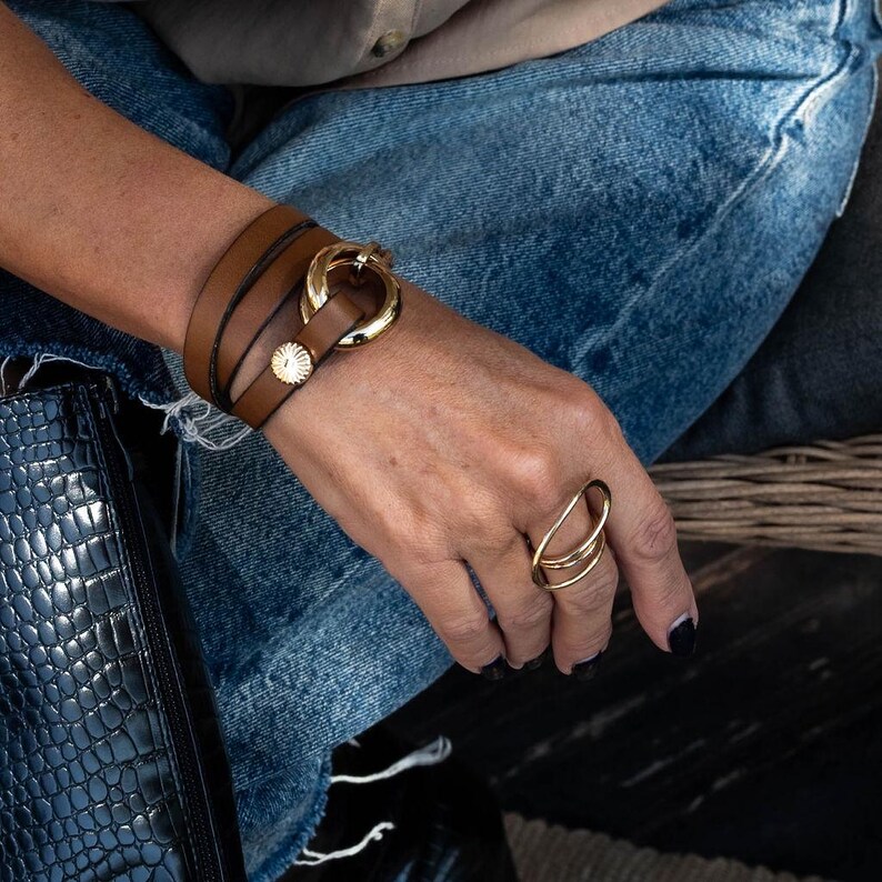 May include: A brown leather wrap bracelet with a gold-colored clasp and a small, round, gold-colored button. A gold-colored ring with a unique, layered design is also visible. The jewelry is worn on a person's wrist and hand.