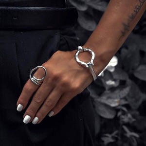Puede incluir: Un conjunto de anillo y pulsera de plata. El anillo presenta un diseño abstracto único con múltiples bandas. La pulsera tiene un estilo similar, con una forma redondeada y abierta y un pequeño detalle de bola. Las joyas se llevan en la mano y la muñeca.