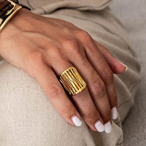 Puede incluir: Un anillo dorado con un diseño rectangular con barras verticales. El anillo se lleva en una mano, con una pulsera dorada visible en el fondo. El diseño del anillo es una característica destacada, que capta la luz y añade un toque de elegancia.