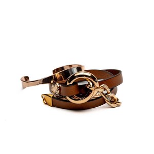 May include: A collection of brown leather bracelets with gold-colored metal accents. One bracelet features a large gold ring, another a gold chain, and a third is a thin, curved cuff. The bracelets are stacked together.