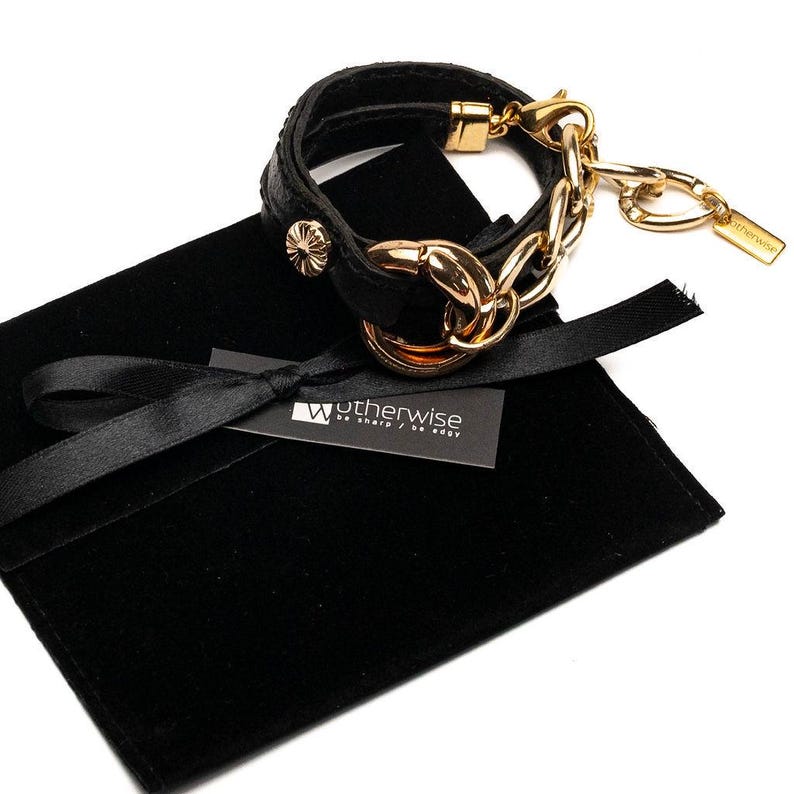 Puede incluir: Pulsera de cuero negro con cadena y herrajes dorados. La pulsera tiene un dise&ntilde;o de doble vuelta con un cierre dorado decorativo y una peque&ntilde;a etiqueta. Presentada en una bolsa de terciopelo negro con una etiqueta "Wotherwise be sharp / be edgy."