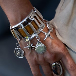 Könnte beinhalten: Ein breites silberfarbenes Manschettenarmband mit brauner, strukturierter Einlage und mehreren hängenden Anhängern, darunter ein Stern, ein kreisförmiges Eulenmedaillon mit griechischen Buchstaben und ein Kreuz. Ein großer, glatter, silberfarbener Ring ist sichtbar.